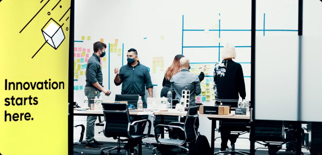 Five people in masks working at a wall of sticky notes in a Digital Surgeons meeting room.