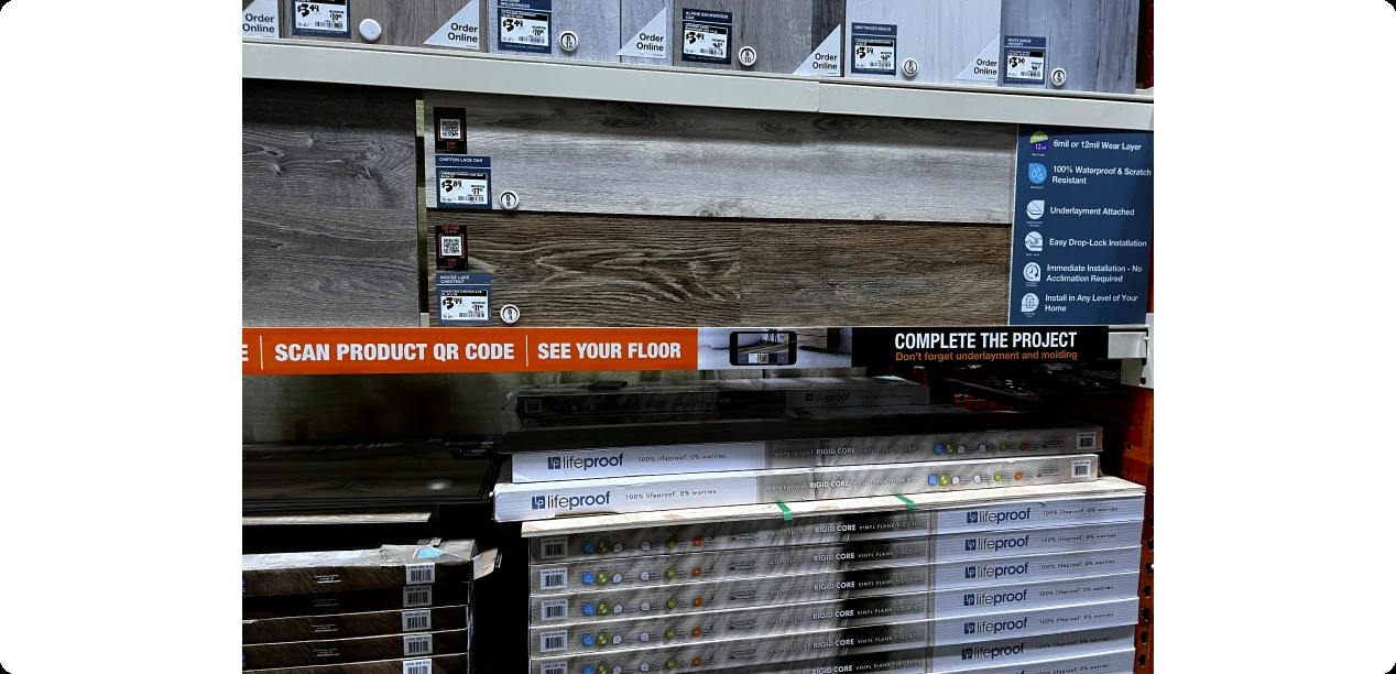 A Home Depot flooring shelf with a sign reading Scan product QR code, See your floor, above stacked flooring boxes.